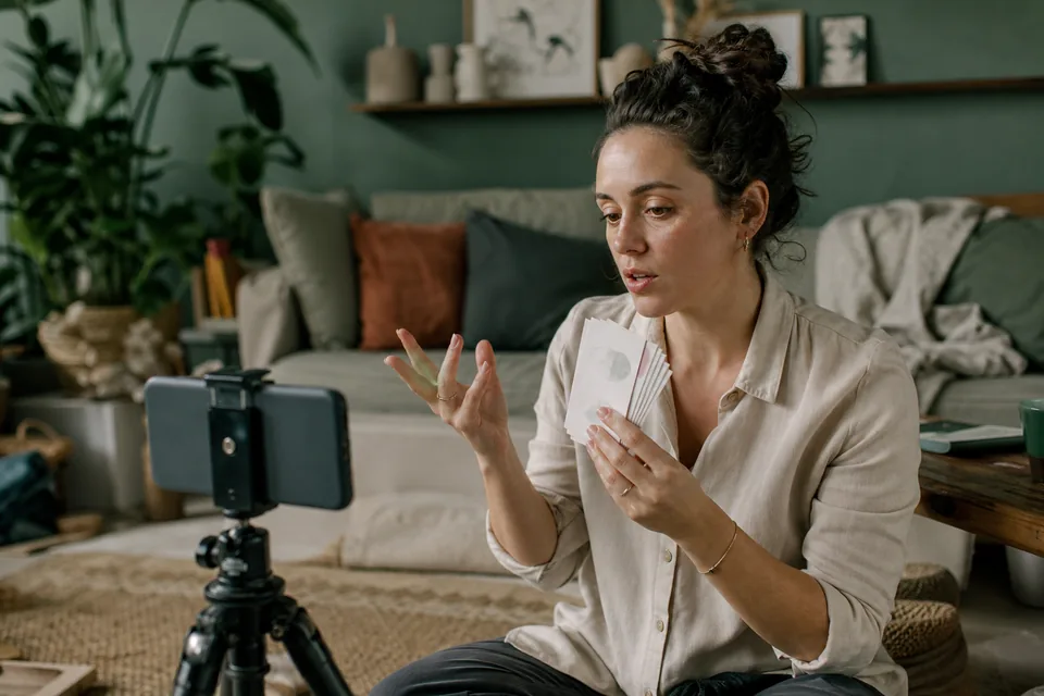 A creator sits on a living room floor and talks to a phone on a tripod, holding a small card in one hand