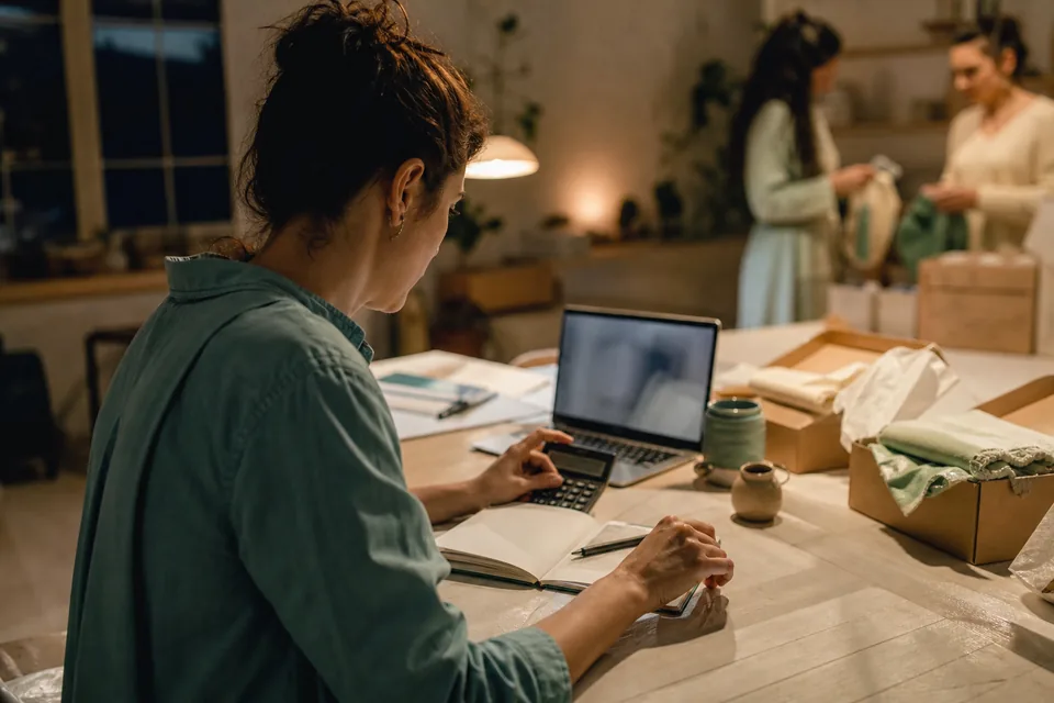 A woman works through numbers on a calculator and laptop at a workshop table, parcels being packed in the background