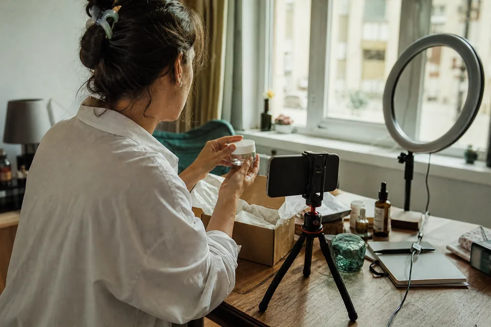 A creator films an unboxing on her phone at a window table, lifting a jar out of kraft packaging