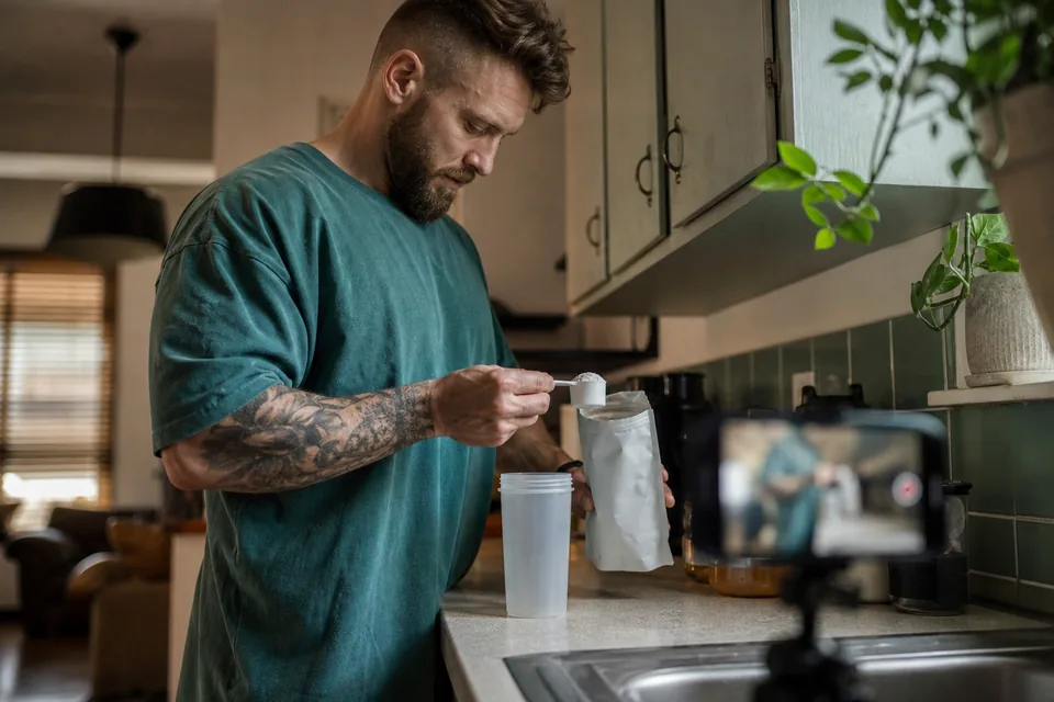 A creator adds a scoop of powder to a smoothie at a kitchen counter in morning light, the label of the tub facing the phone camera